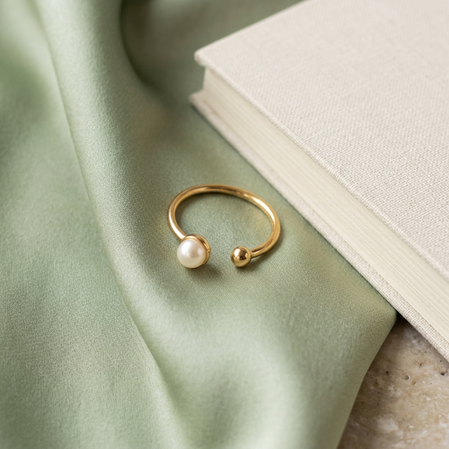 Pearl-style gold-tone open ring on pale sage satin beside a cream notebook